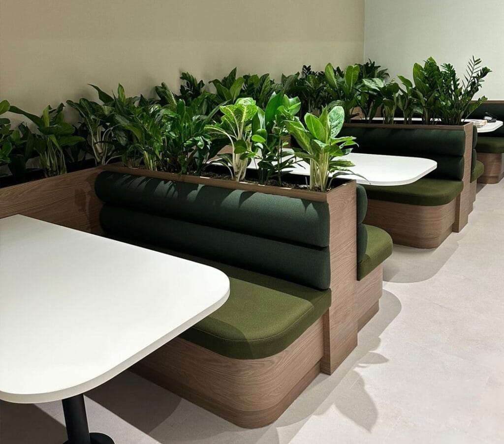 Modern dining booths with green upholstery and integrated planters filled with lush green plants, in an office or commercial setting.
